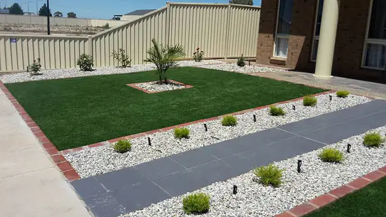 Front yard with a lawn, white pebble garden beds, small shrubs and a grey paved path