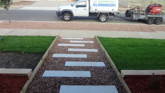 Stepping-stone path through pebbles between two new lawns, with a Goulburn Valley Sprinklers ute and trailer parked in the street