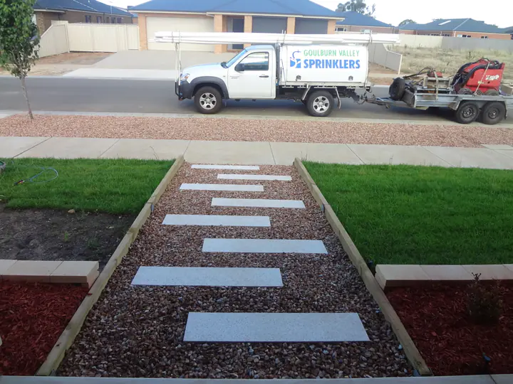 Stepping-stone path through pebbles between two new lawns, with a Goulburn Valley Sprinklers ute and trailer parked in the street